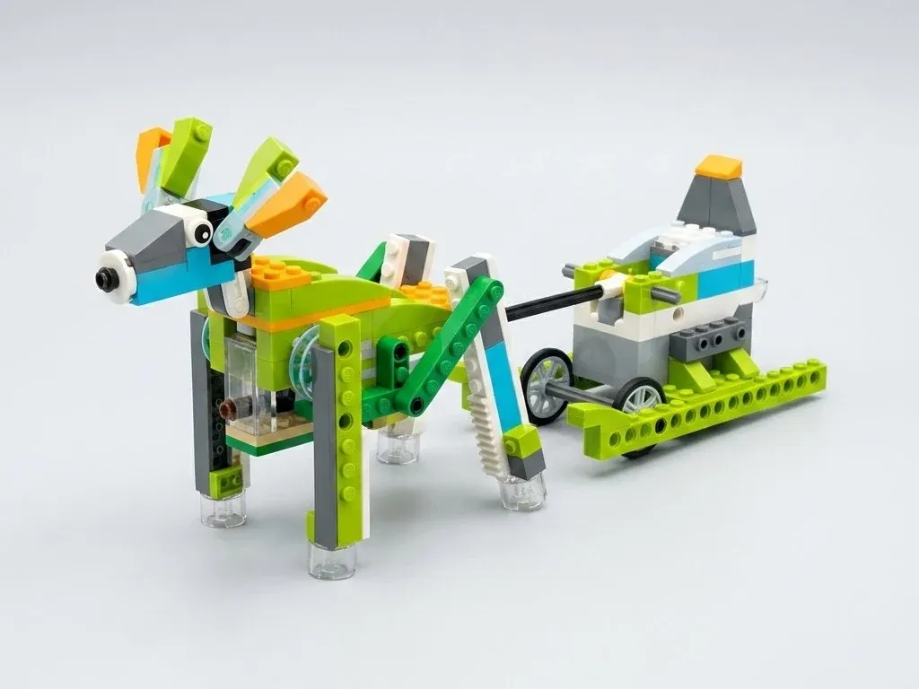 Олень Рудольф из деталей, совместимых с WeDo 2.0, с оранжево-зелёными рогами шагает на четырёх ногах и тянет за чёрную ось сани; на боку виден голубой диск с чёрным штифтом у края и зелёная тяга к ноге